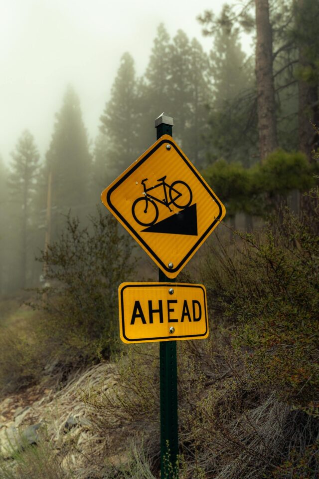 Żółty rowerowy znak ostrzegawczy na zamglonej leśnej ścieżce, wskazujący zjazd w dół.